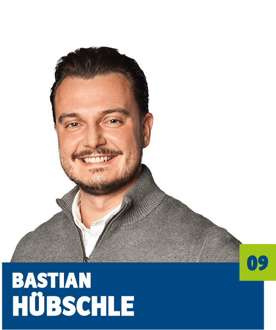 Professionelles Porträt von Bastian Hübschle vor dunklem Hintergrund. Er trägt ein helles Hemd und einen grauen Pullover mit Reißverschluss und lächelt freundlich in die Kamera. Der offene Blick und das gepflegte Erscheinungsbild vermitteln Kompetenz, Sympathie und eine moderne, vertrauensvolle Ausstrahlung und eignen sich sehr gut für die persönliche Vorstellung im beruflichen oder organisatorischen Kontext.