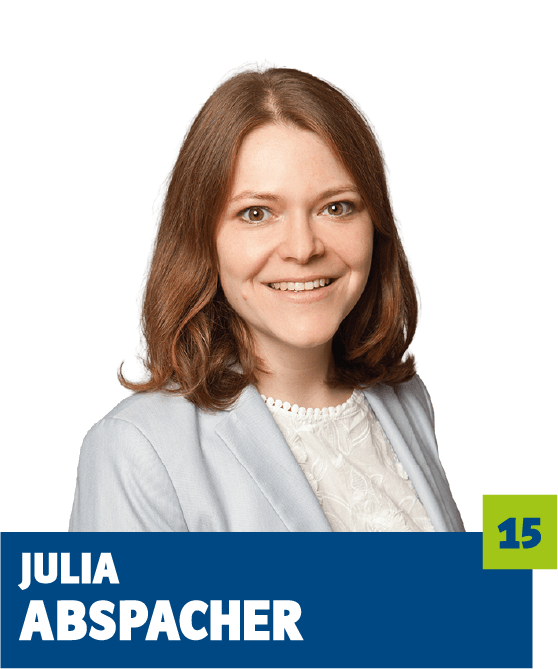 Professionelles Porträt von Julia Abspacher vor dunklem Hintergrund. Sie trägt eine helle Bluse und ein hellblaues Sakko und lächelt freundlich in die Kamera. Der offene Blick und die harmonische Farbwahl vermitteln Kompetenz, Frische und eine sympathische, vertrauensvolle Ausstrahlung und eignen sich ideal für die persönliche Vorstellung im beruflichen oder organisatorischen Kontext.