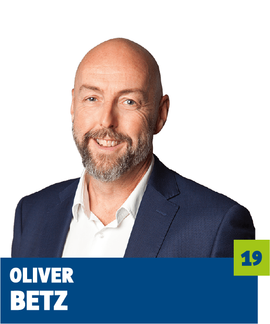 Professionelles Porträt von Oliver Betz vor dunklem Hintergrund. Er trägt ein weißes Hemd und ein dunkles Sakko und lächelt freundlich in die Kamera. Der offene Blick, der gepflegte Bart und das souveräne Auftreten vermitteln Kompetenz, Seriosität und Vertrauen und eignen sich sehr gut für die persönliche Vorstellung im beruflichen oder organisatorischen Kontext.