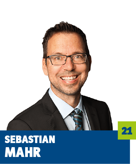 Professionelles Porträt von Sebastian Mahr vor dunklem Hintergrund. Er trägt ein helles Hemd, eine Krawatte, ein dunkles Sakko sowie eine Brille und lächelt offen in die Kamera. Der freundliche Gesichtsausdruck und der direkte Blick vermitteln Kompetenz, Seriosität und eine positive, vertrauensvolle Ausstrahlung und eignen sich ideal für die persönliche Vorstellung im beruflichen oder organisatorischen Kontext.