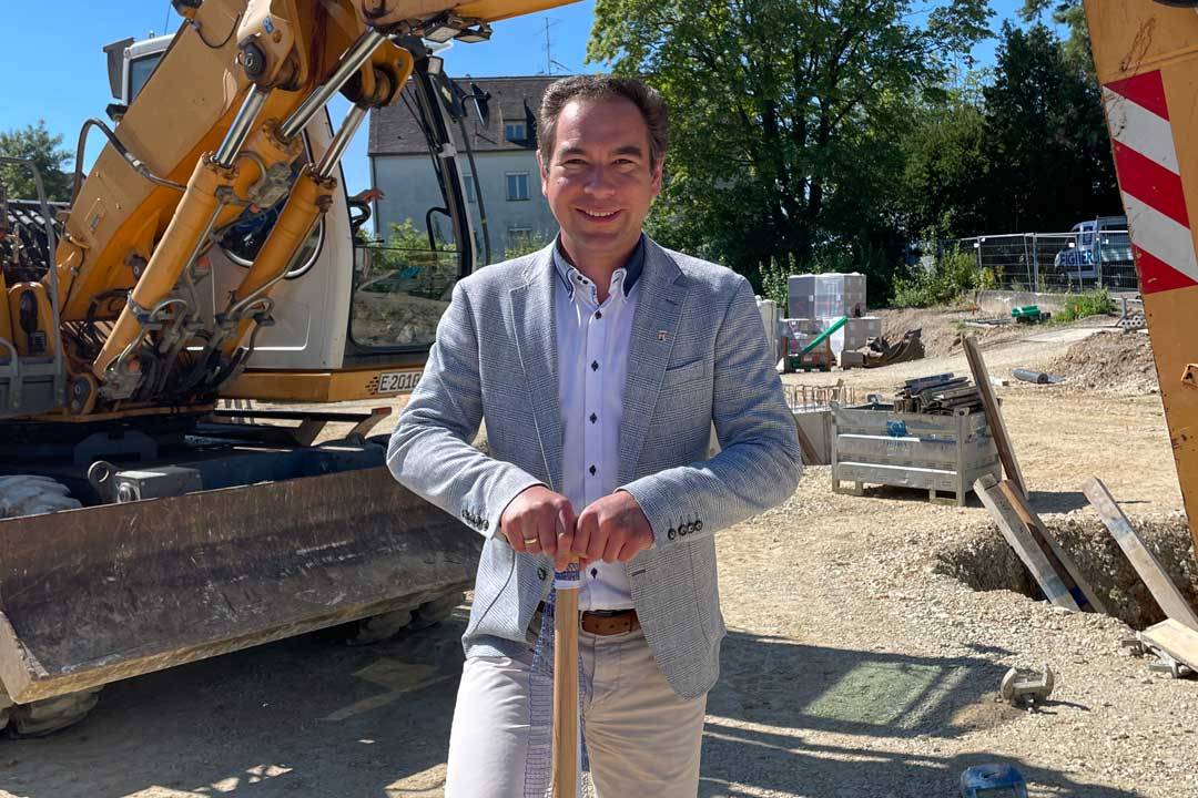 Eine Person steht lächelnd auf einer Baustelle und hält einen Spaten vor sich. Sie trägt ein helles Hemd, ein Sakko und eine helle Hose und wirkt gepflegt und präsent. Im Hintergrund sind ein Bagger, Baumaterialien und eine laufende Baustelle zu sehen. Die Szene verbindet persönlichen Einsatz mit einem baulichen Umfeld und vermittelt Engagement, Nähe zur Praxis und einen aktiven Bezug zu einem Bau- oder Infrastrukturprojekt.