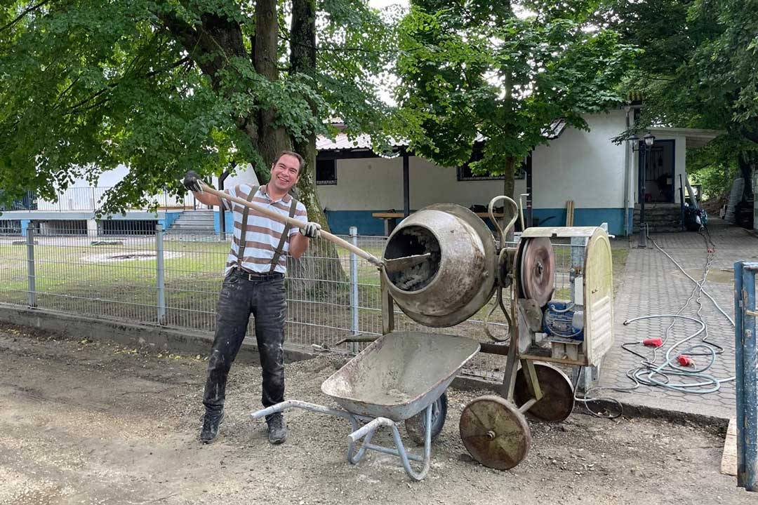Eine Person steht im Freien auf einer Baustelle und arbeitet mit einem laufenden Betonmischer. Sie hält eine Schaufel in den Händen und füllt frischen Beton in eine Schubkarre. Die Kleidung ist praktisch und sichtbar verschmutzt, was auf aktive Handarbeit hindeutet. Im Hintergrund sind Bäume, ein Zaun sowie Gebäude zu sehen. Die Szene vermittelt Tatkraft, handwerklichen Einsatz und eine bodenständige, praktische Arbeitssituation im Außenbereich.