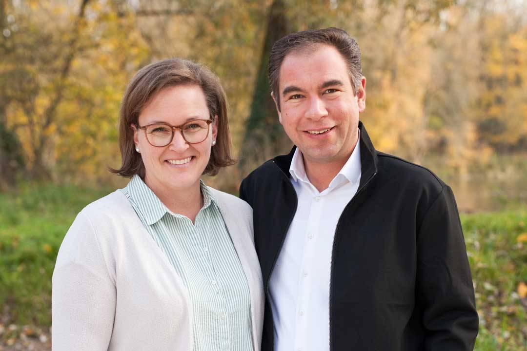 Zwei Personen stehen nebeneinander im Freien und lächeln freundlich in die Kamera. Sie tragen helle, gepflegte Alltagskleidung und wirken entspannt und nahbar. Im Hintergrund sind Bäume mit herbstlicher Laubfärbung und eine grüne Wiese zu sehen, was der Aufnahme eine warme, natürliche Atmosphäre verleiht. Das Bild vermittelt Nähe, Vertrauen und einen persönlichen Moment in ruhiger Umgebung.