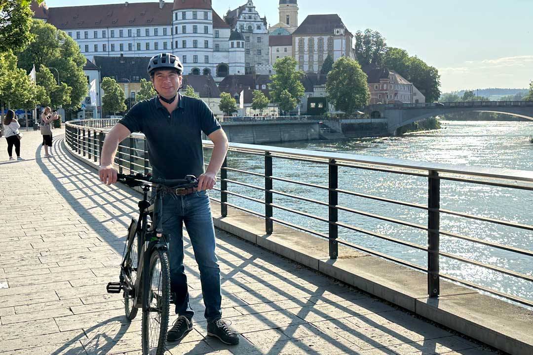Eine Person steht mit einem Fahrrad auf einer gepflasterten Uferpromenade und blickt lächelnd in die Kamera. Sie trägt einen Fahrradhelm, ein dunkles Poloshirt und Jeans. Im Hintergrund sind ein Fluss, eine Brücke sowie eine historische Altstadt mit markanten Gebäuden und Türmen zu sehen. Die Aufnahme vermittelt Aktivität, Lebensfreude und Verbundenheit mit der Stadt und ihrer Umgebung.
