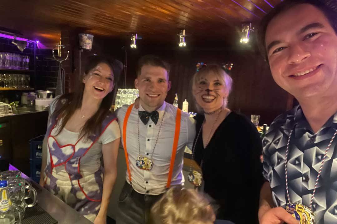 Vier Personen stehen gut gelaunt hinter einer Bar und lächeln in die Kamera. Sie tragen festliche Kostüme und Accessoires, darunter Hosenträger, Fliege, Orden sowie geschminkte Gesichter, was auf eine Faschings- oder Karnevalsveranstaltung hindeutet. Die Bar im Hintergrund ist beleuchtet und mit Gläsern und Getränken ausgestattet. Die Szene wirkt ausgelassen, herzlich und vermittelt Gemeinschaft, Feierlaune und einen fröhlichen Moment in geselliger Atmosphäre.