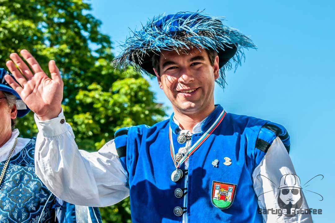 Eine Person trägt eine auffällige blaue Festtracht mit weißem Hemd und verziertem Wams sowie einen passenden Hut mit Federbesatz. Sie hebt freundlich die Hand zum Gruß und lächelt in die Kamera. An der Kleidung sind Abzeichen und ein Wappen angebracht, was auf eine traditionelle oder historische Veranstaltung hindeutet. Der Hintergrund mit blauem Himmel und grünen Bäumen unterstreicht die festliche, lebendige Atmosphäre des Moments.