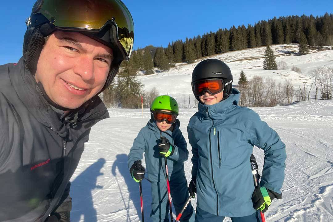 Ein Mann und zwei Kinder stehen auf einem verschneiten Skigelände und posieren für ein Selfie. Alle tragen Skiausrüstung, inklusive Helmen und Brillen, und sind in warmen Outdoor-Kleidern gekleidet. Der Mann lächelt in die Kamera, während die Kinder ihm gegenüber ebenfalls einen freundlichen Ausdruck zeigen. Im Hintergrund ist eine verschneite Landschaft mit Bäumen und Hügeln zu sehen, was das winterliche Skierlebnis unterstreicht.