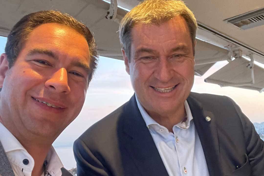 Zwei Männer stehen nah beieinander und lächeln in die Kamera. Beide tragen helle Hemden und Sakkos und wirken gepflegt und gut gelaunt. Die Aufnahme entstand offenbar im Innenbereich mit Blick nach draußen, möglicherweise in einem Café oder Veranstaltungsraum mit Überdachung. Das Bild vermittelt Nähe, Austausch und eine positive, persönliche Atmosphäre.