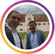 Zwei Personen stehen im Freien und blicken lächelnd in die Kamera. Im Hintergrund ist ein historisches Gebäude mit hellem Putz und markantem Dach zu sehen, umgeben von einer Grünfläche. Die Aufnahme wirkt ungezwungen und persönlich und vermittelt Nähe, Austausch und einen informellen Moment in einem repräsentativen Umfeld.