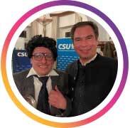Zwei Personen stehen nebeneinander und lächeln in die Kamera. Eine Person trägt einen Anzug und zeigt mit erhobenem Daumen eine positive Geste, die andere trägt ein dunkles Hemd. Im Hintergrund sind mehrfach CSU-Logos zu sehen, was auf eine politische Veranstaltung oder ein Treffen im parteinahen Umfeld hinweist. Das Bild vermittelt Offenheit, Verbundenheit und eine positive Stimmung in einem politischen Kontext.