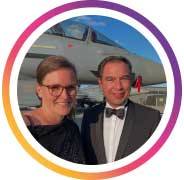 Zwei Personen stehen nebeneinander und lächeln in die Kamera. Sie tragen festliche Kleidung, darunter ein Anzug mit Fliege und ein elegantes Outfit, und posieren vor einem Flugzeug im Hintergrund. Die Aufnahme wirkt persönlich und feierlich und vermittelt Nähe, Freude sowie seehr einen besonderen Anlass in stilvollem Umfeld.