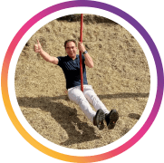 Eine Person schaukelt im Freien über trockenem Gras und hält sich mit beiden Händen an den Seilen der Schaukel fest. Sie trägt helle Kleidung und lächelt entspannt, während sie sich leicht nach hinten lehnt. Die Szene wirkt locker, ungezwungen und vermittelt Freude, Bewegung und eine persönliche, lebensnahe Atmosphäre.